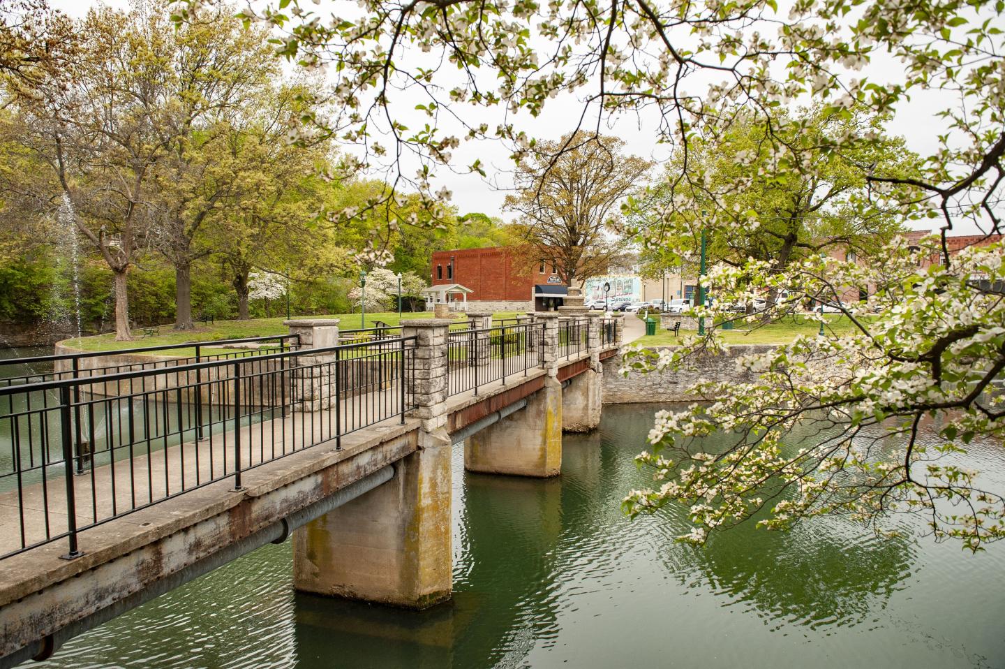 Bridge over a green river, surrounded by trees and a red building in the background.