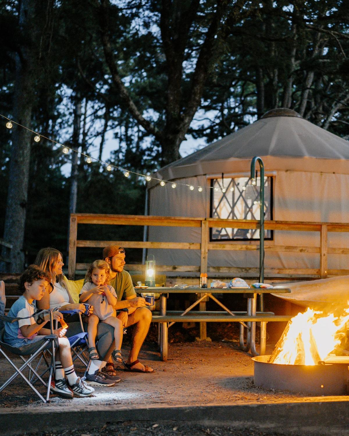 Family sitting by a campfire near a yurt, surrounded by string lights at dusk.