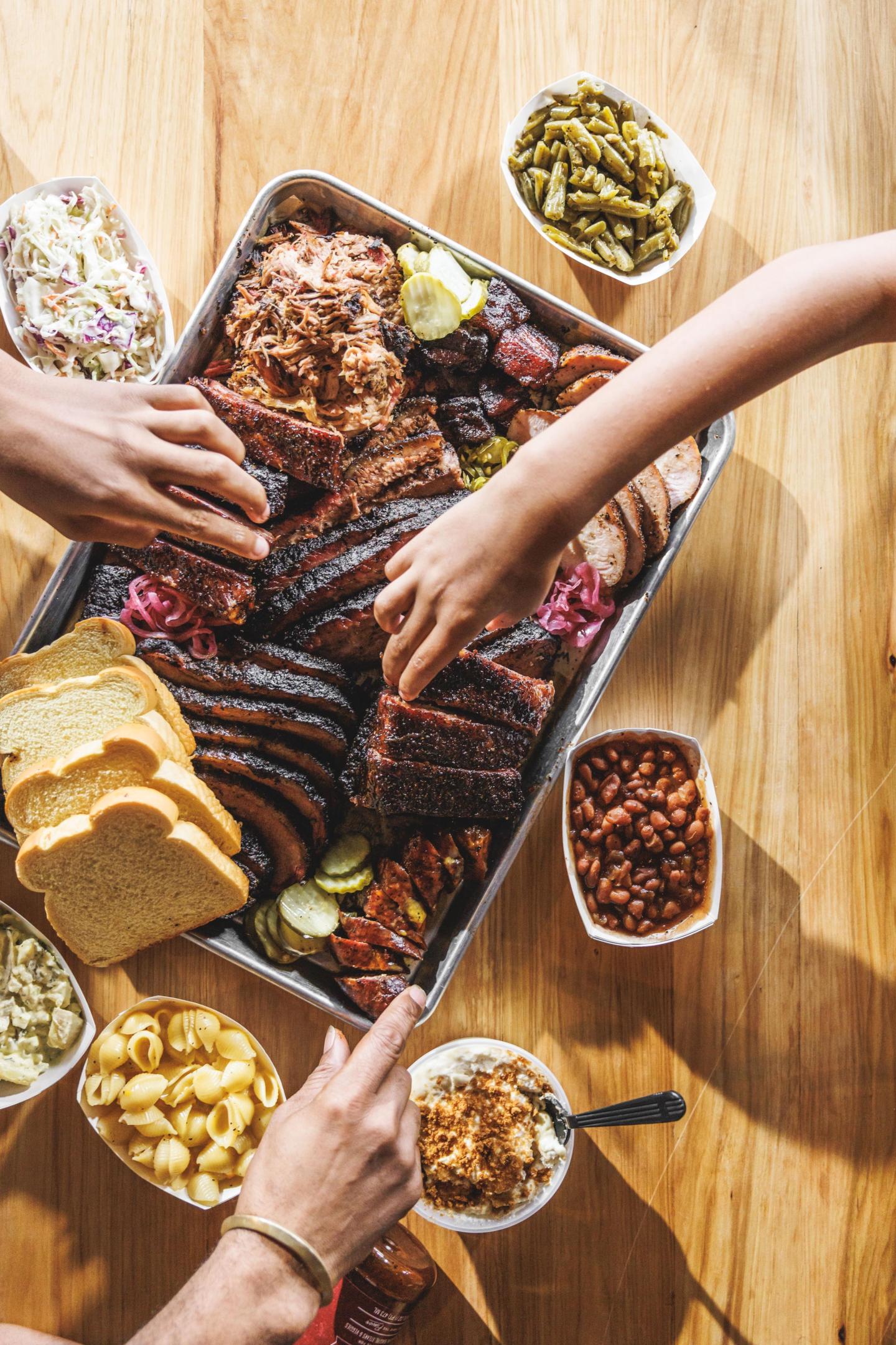Hands reaching for barbecue platter with sides on wooden table.