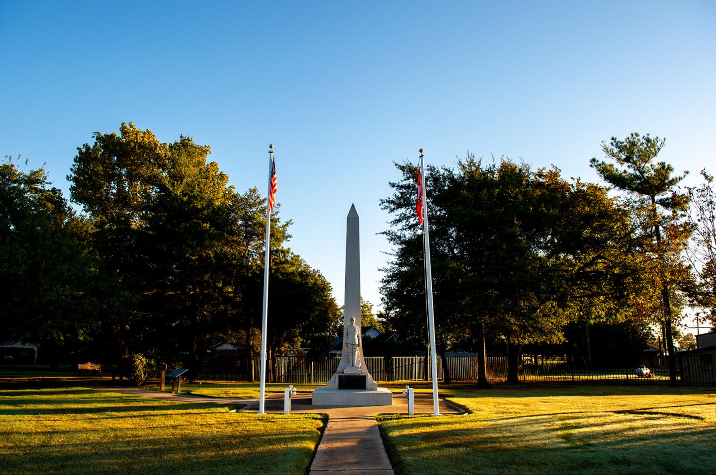 Tall stone monument between two flagpoles in a park with green grass and trees.