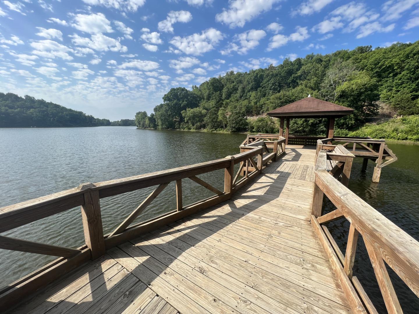 Wooden pier leads to gazebo on calm lake, surrounded by lush trees under blue sky.