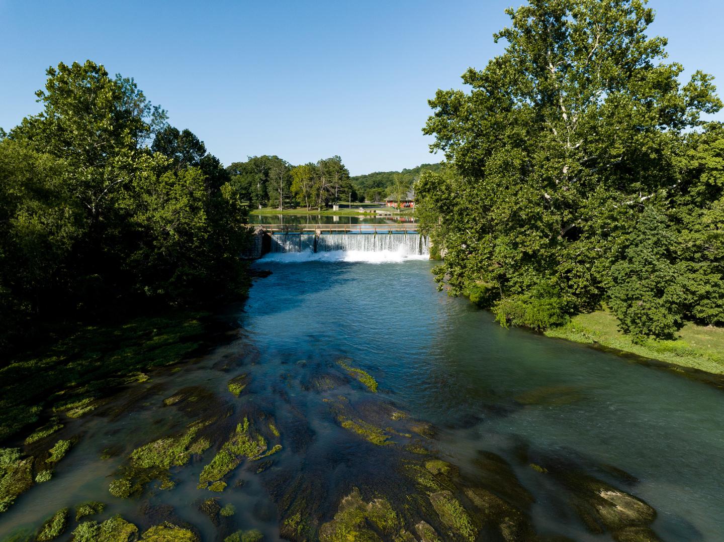 River flowing under a bright sky, bordered by lush trees, with a small dam waterfall in the distance.