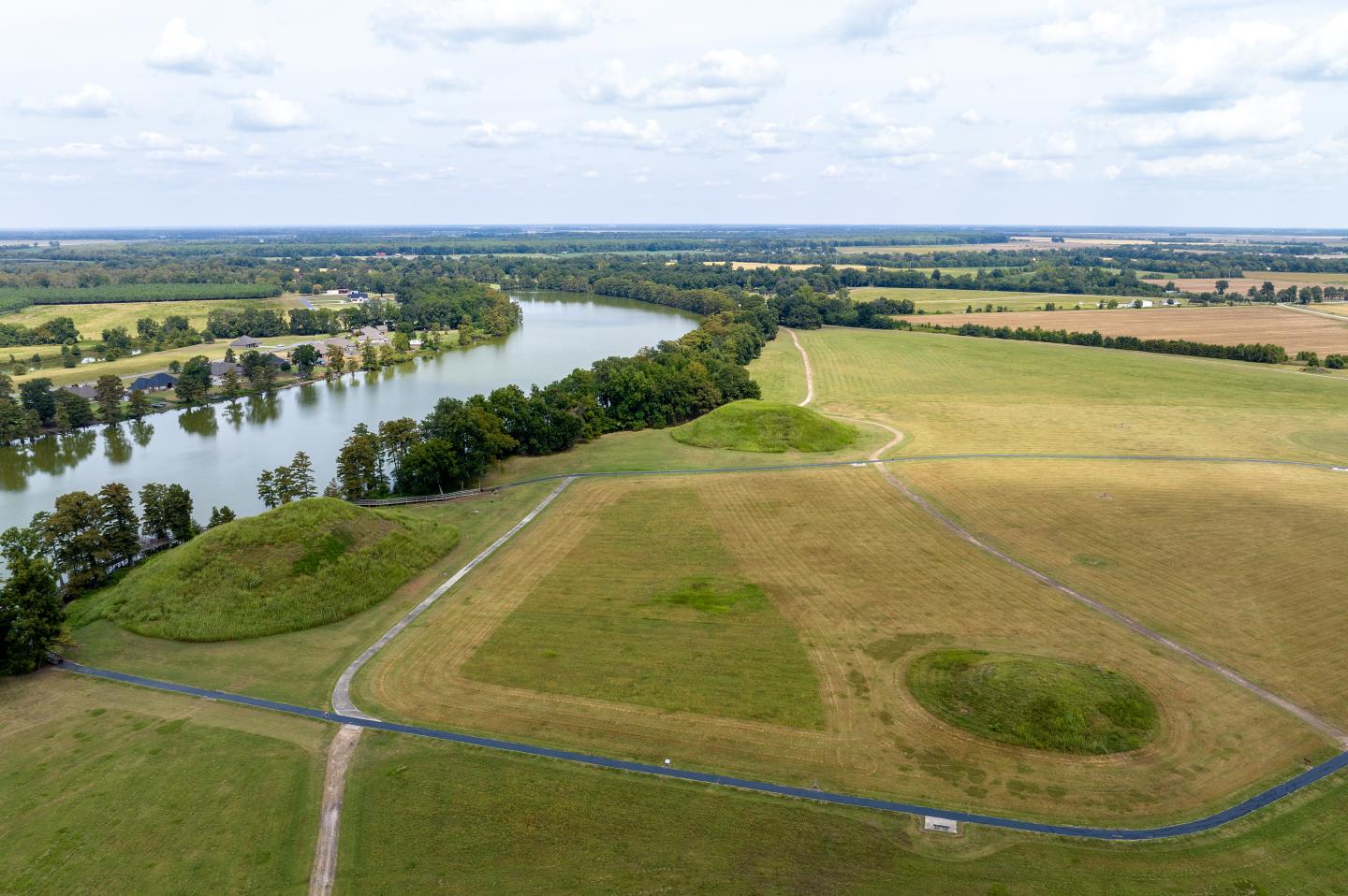 Aerial view of a grassy field by a river, under a blue sky with clouds.