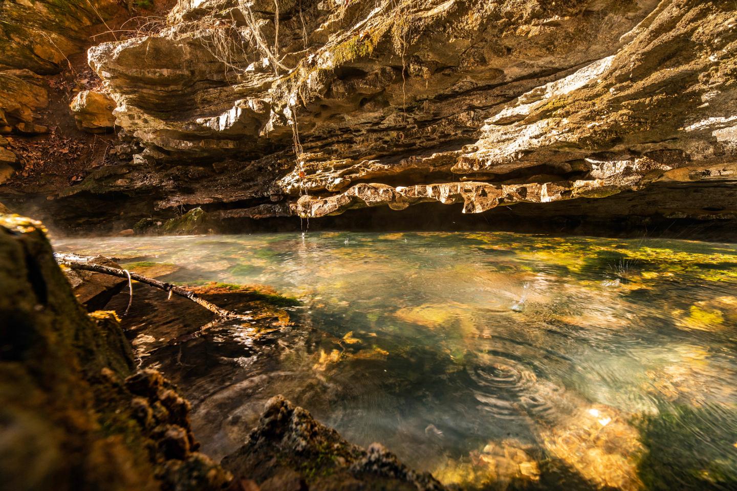 Rocky cave with a reflective pool of water, illuminated by sunlight.