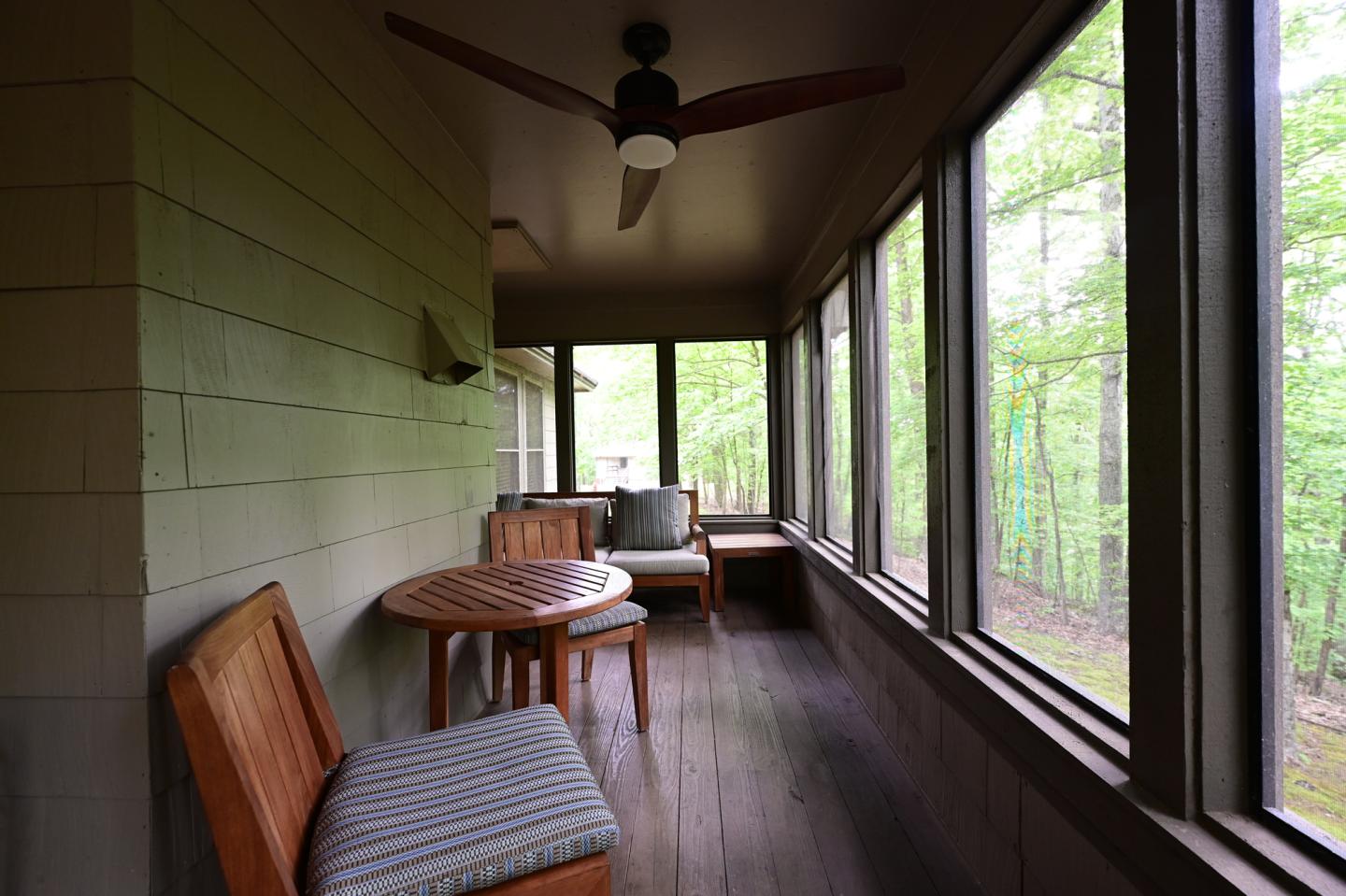 Screened porch with wooden chairs, round table, ceiling fan, and forest view.