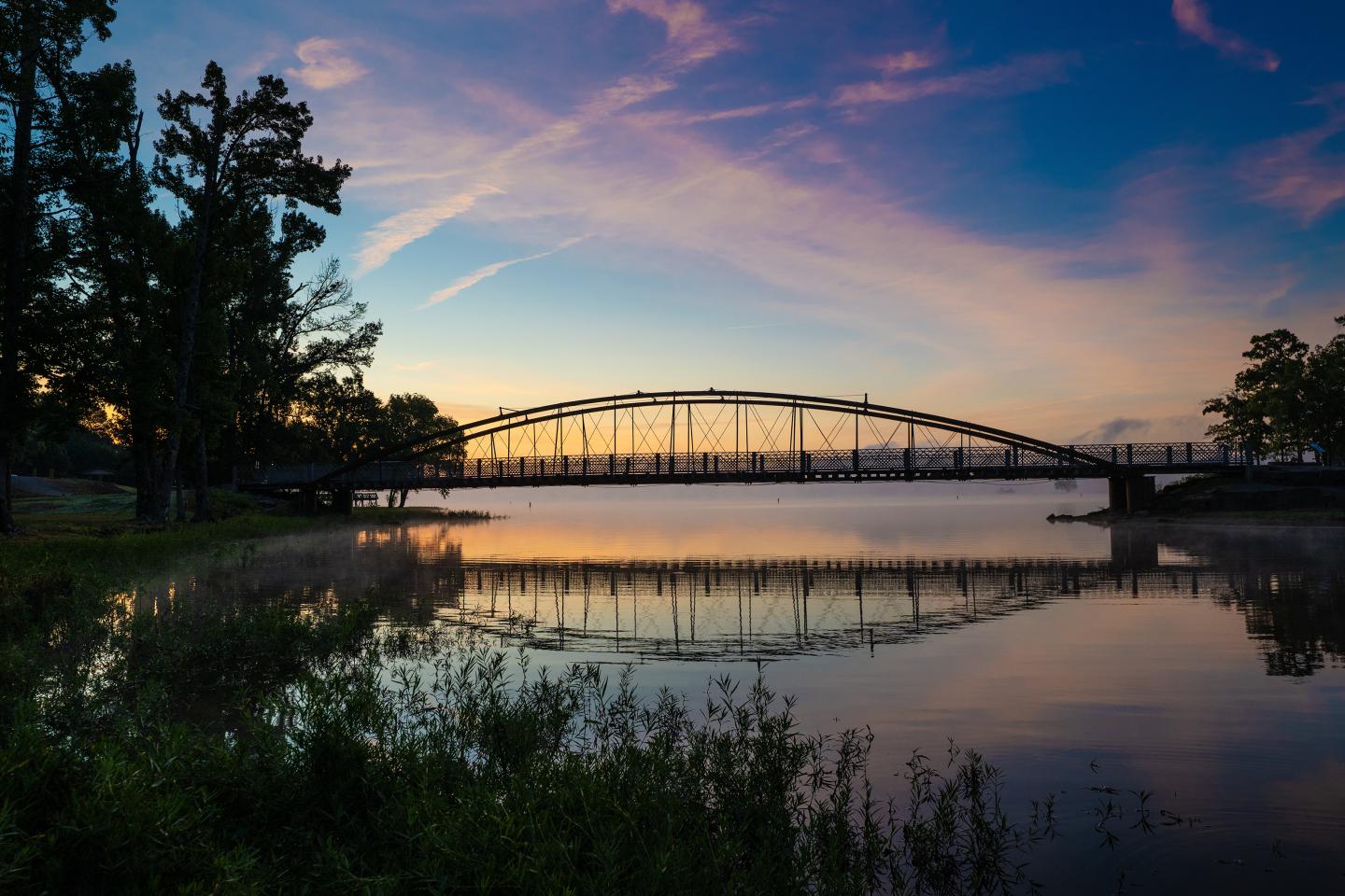 Bridge over calm water at sunset, with trees silhouetted against a colorful sky.