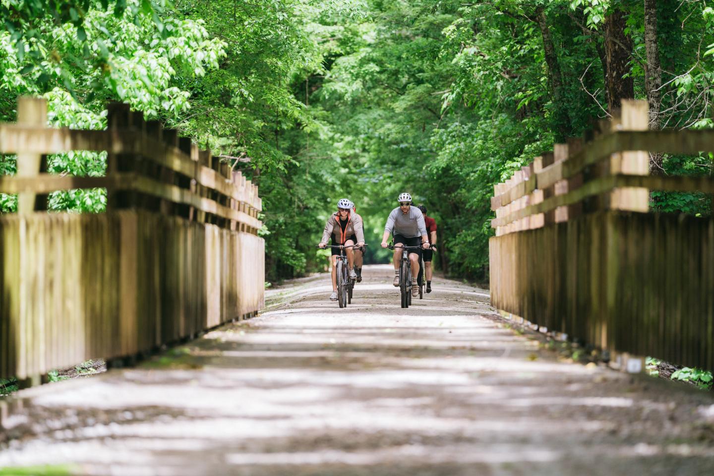 Two cyclists ride on a gravel path with wooden fencing in a green forest.