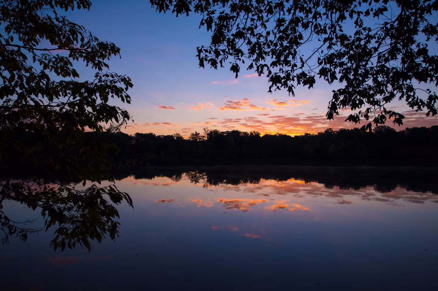 Twilight over a calm lake with silhouetted trees and colorful sky reflections.
