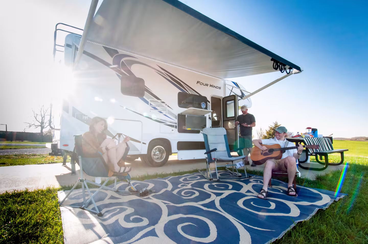 RV parked with people relaxing outside; one plays guitar under the sunny sky.