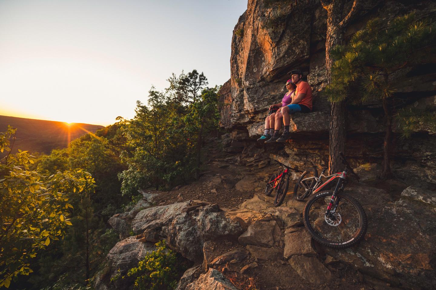 Two people sitting on a cliff, with bikes, at sunset.