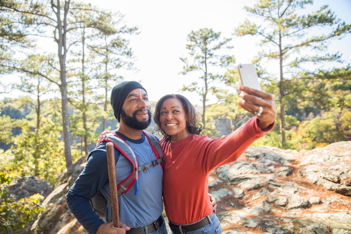 Couple takes a selfie while hiking, bright day, trees in background.