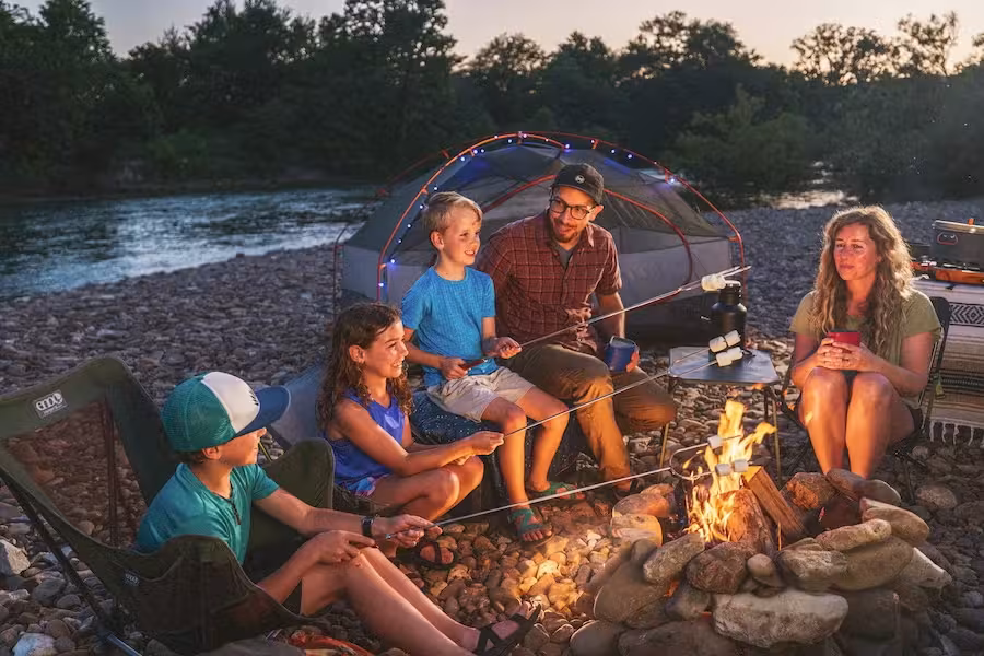 Family camping by a river, sitting around a campfire with a tent in the background.