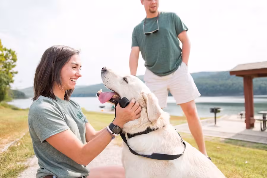 Woman smiling at a happy dog by a lakeside, man in background.