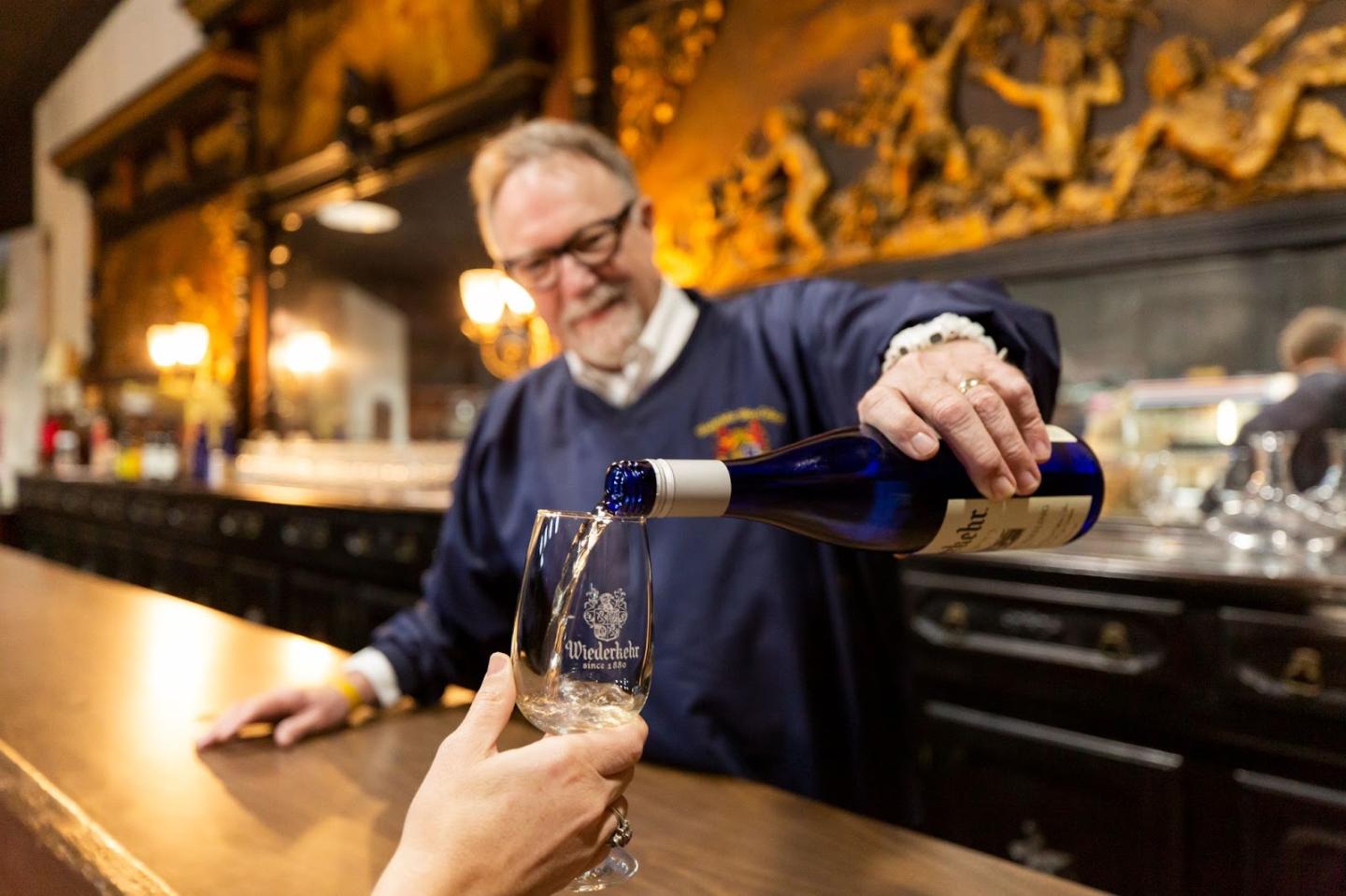 Bartender pouring white wine into a glass at a bar with ornate decor.