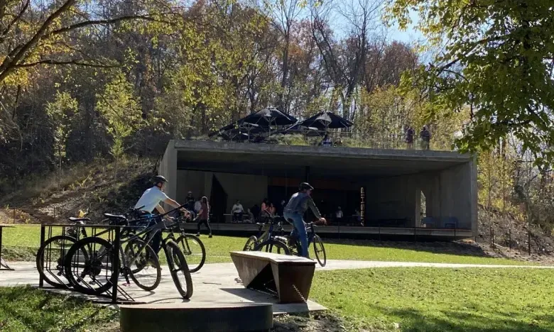Concrete structure in park with people biking nearby, surrounded by trees.