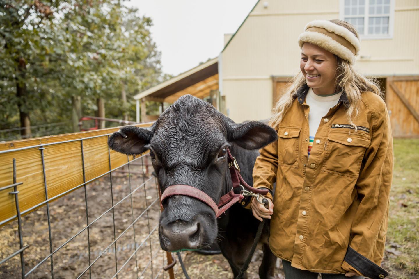 Woman in a brown jacket smiles beside a black cow in a rural setting.