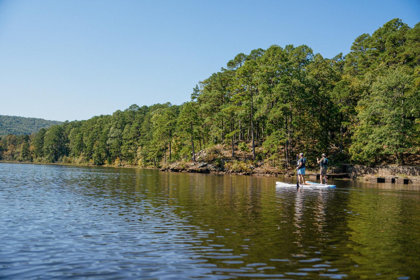 Two people paddleboarding on a calm lake beside a forested shoreline.