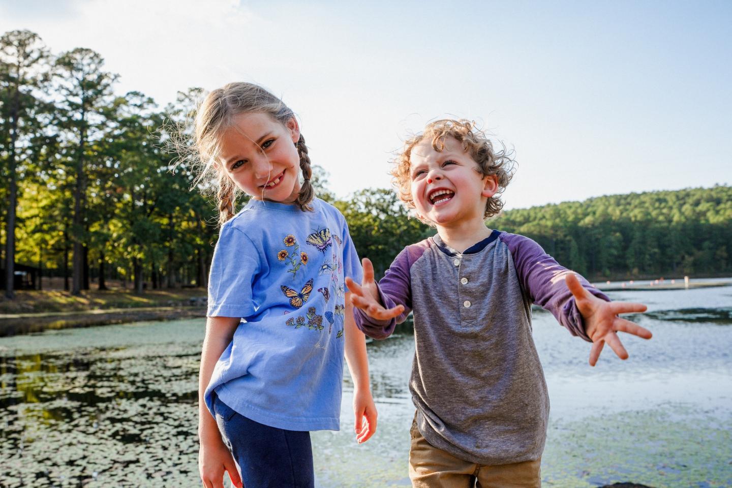 Children laughing by a pond with trees in the background.