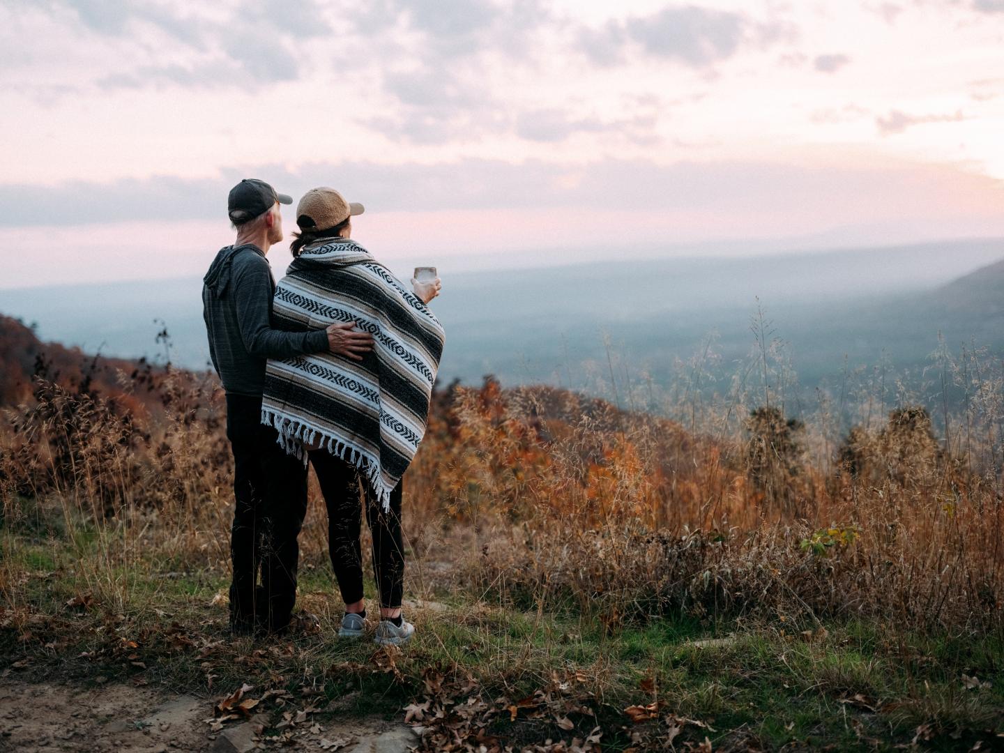 Couple stands on a hill, wrapped in a blanket, watching a scenic sunset.