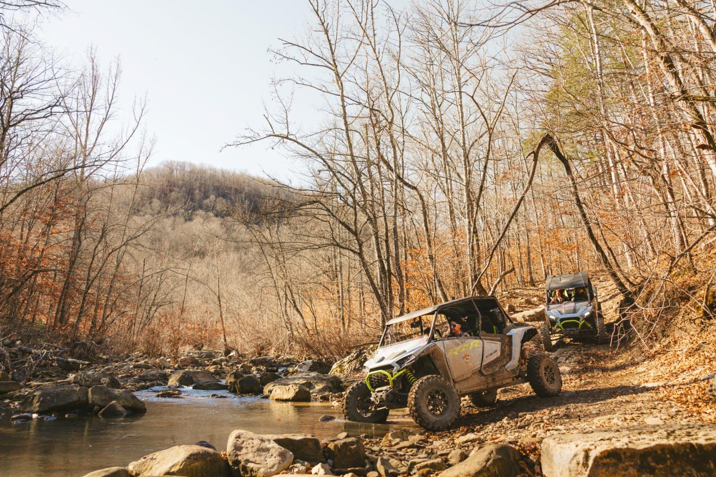 Two off-road vehicles driving beside a rocky stream in a forested area.