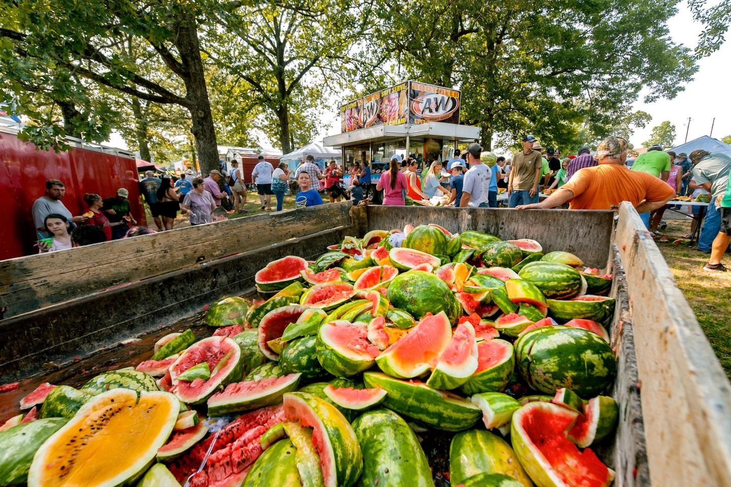 Watermelons in a wooden cart at an outdoor festival with people in the background.