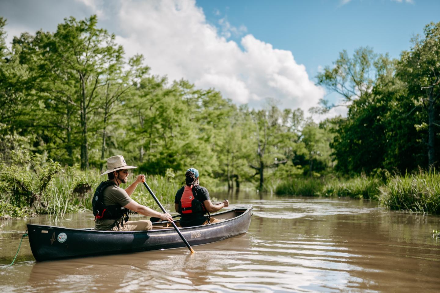 Two people canoeing on a calm river, surrounded by lush greenery and a blue sky.