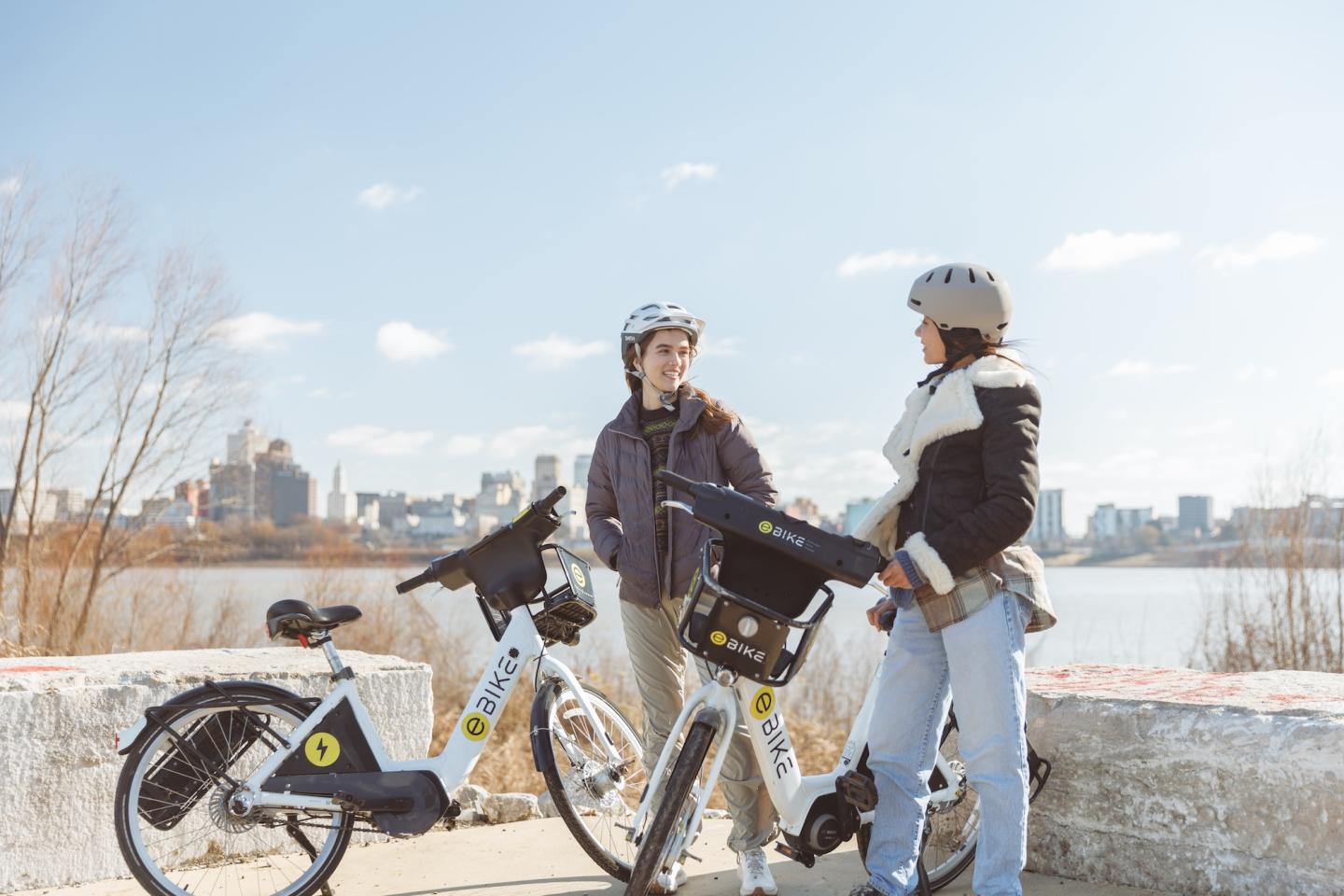 Two people with helmets and bikes, talking by a river on a sunny day. City skyline in background.
