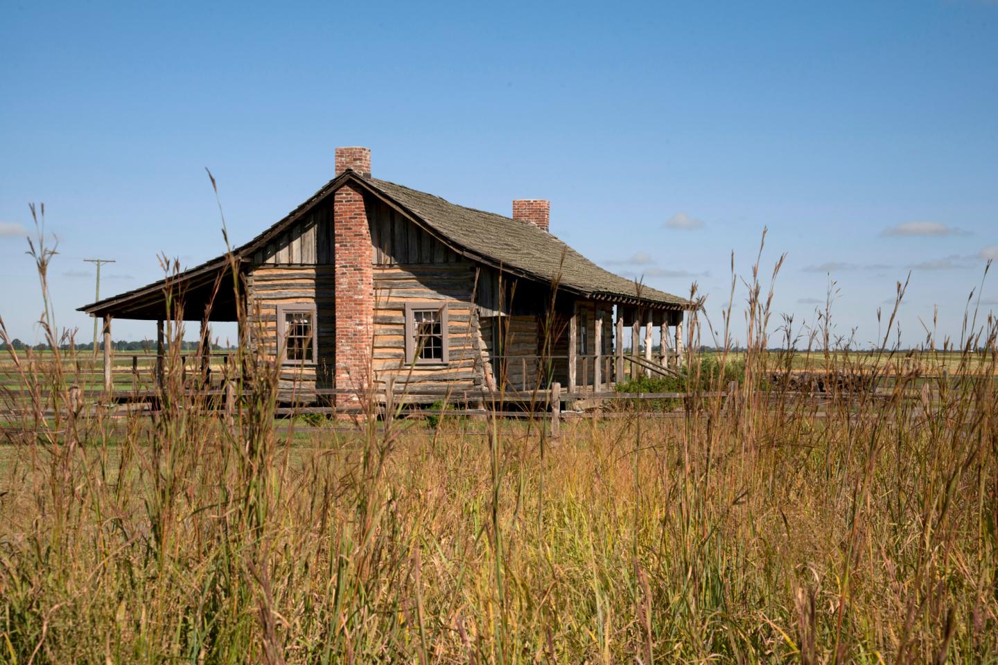 Rustic wooden cabin in a grassy field under a clear blue sky.