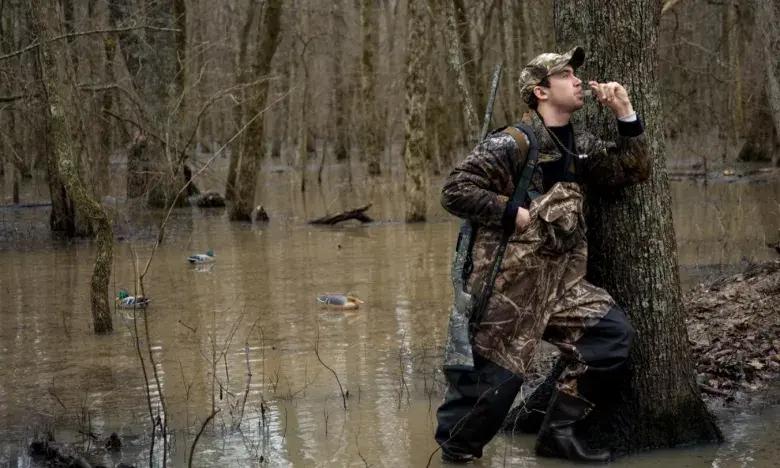Hunter in camo gear using a duck call in a flooded forest.