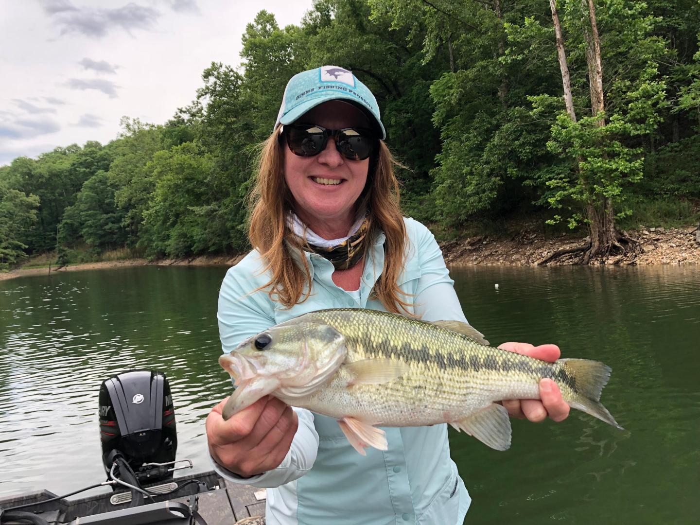 Smiling person holding a fish on a boat in a wooded lake.