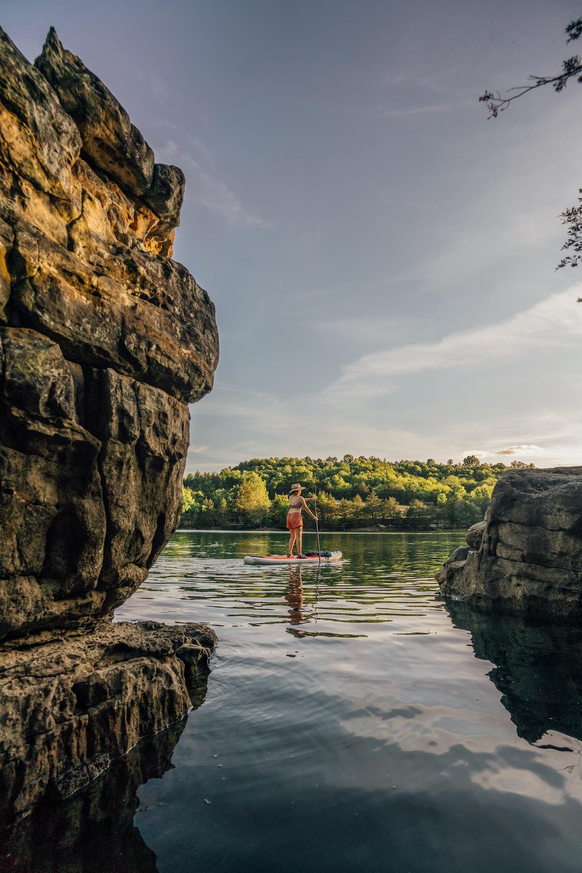 Paddleboarder on calm lake surrounded by cliffs and trees.