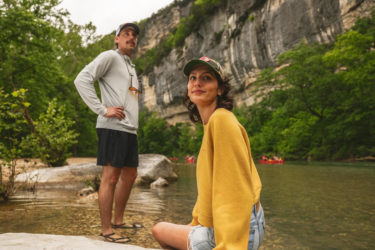 Two people by a river, surrounded by green trees and a rocky cliff.