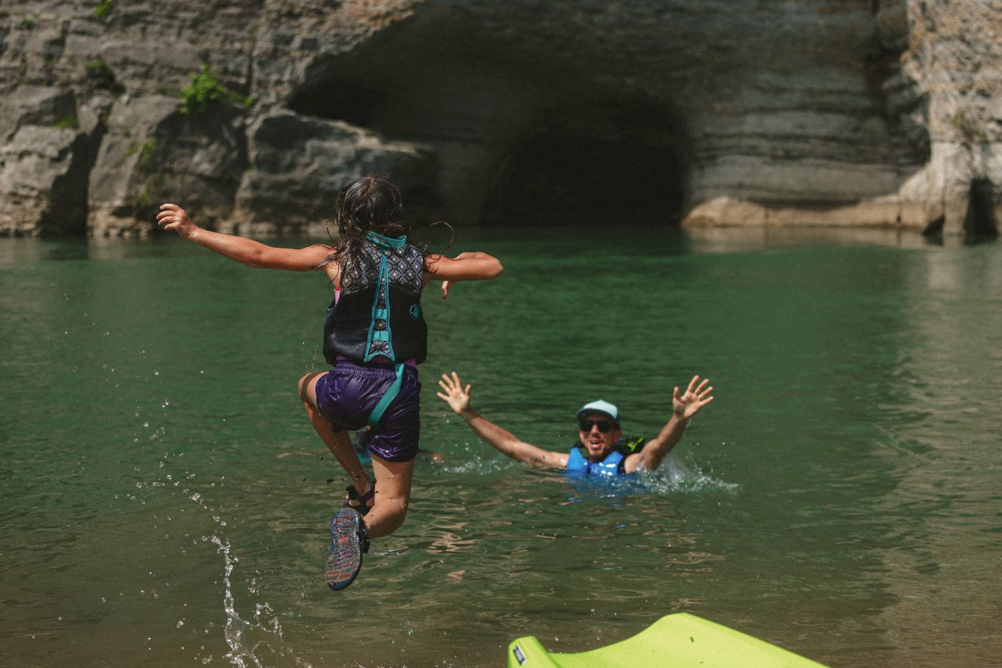 Child jumping into a river towards an adult with arms open, cave in the background.