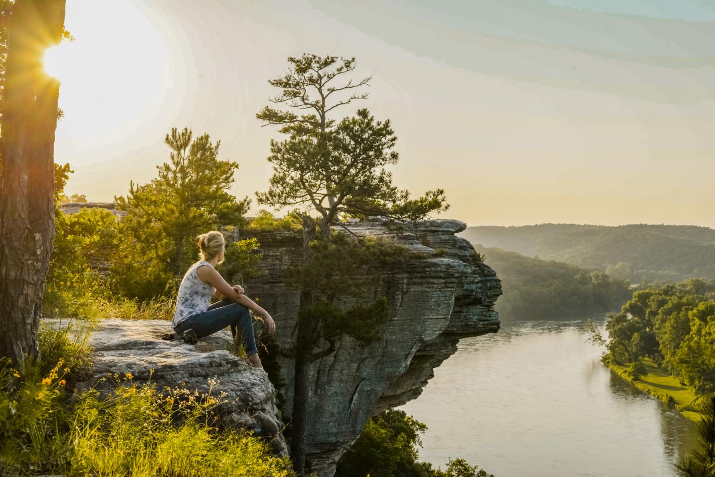 Person sitting on a cliff overlooking a river at sunset.