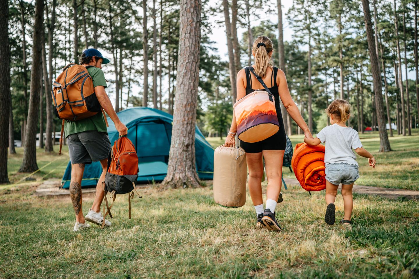 Family carrying camping gear towards tent in a forest.