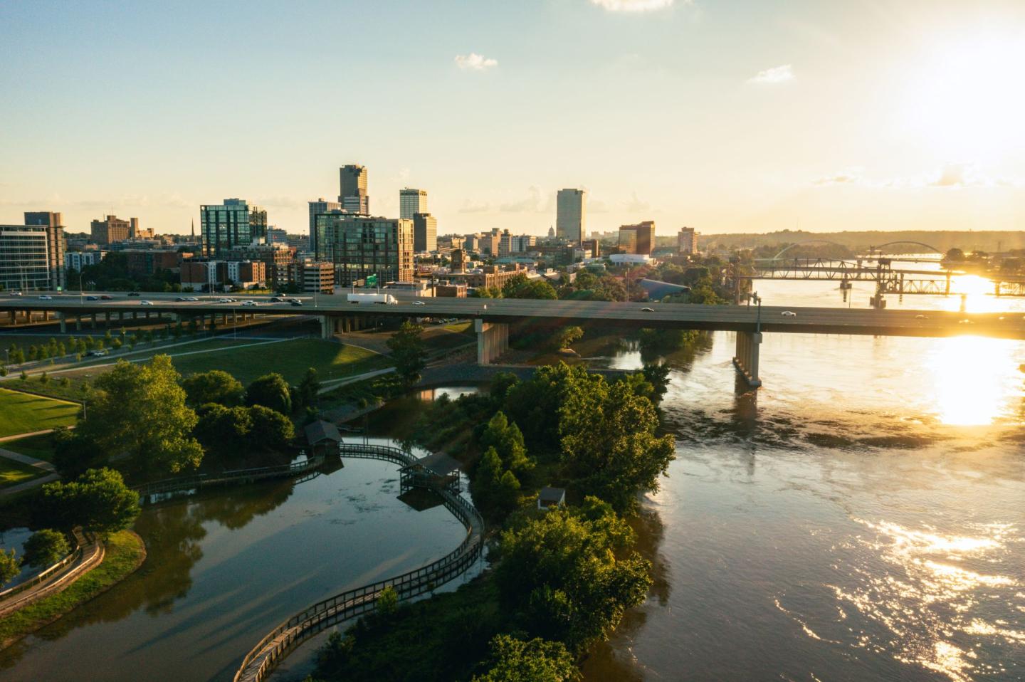 City skyline with a bridge over a river at sunset, greenery in the foreground.
