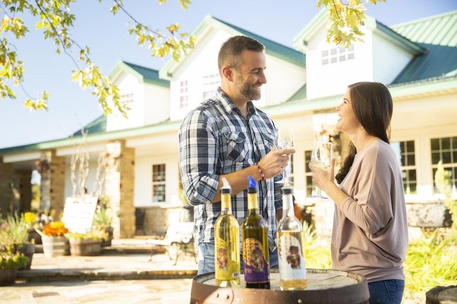 Man and woman clinking glasses outside a house with wine bottles on a barrel.