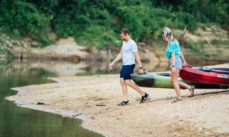 Couple carrying kayaks to the water on a sandy riverbank.