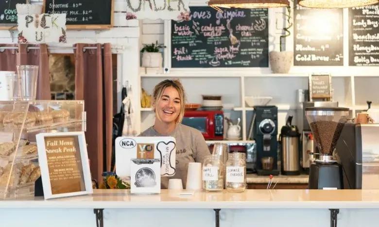 Barista smiling behind a café counter with a coffee machine and menu boards.