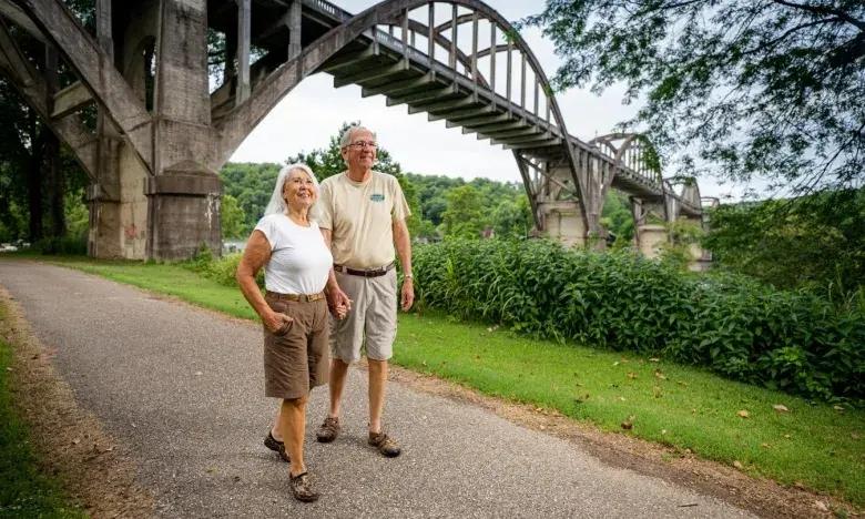 Older couple walking on a park path under a large arch bridge, surrounded by greenery.