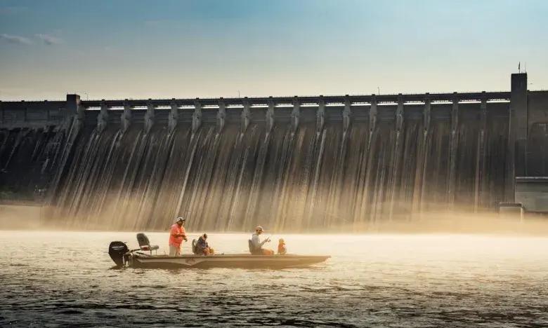 Boat with people fishing near a large dam at sunrise.
