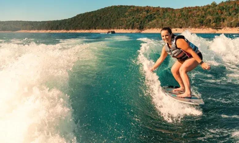 Person wakesurfing on a sunny lake with forested hills in background.