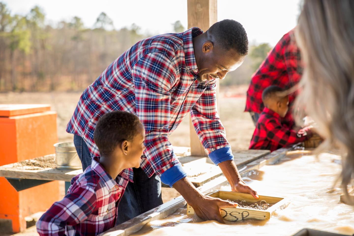 Man and child in matching plaid shirts making pottery outdoors.