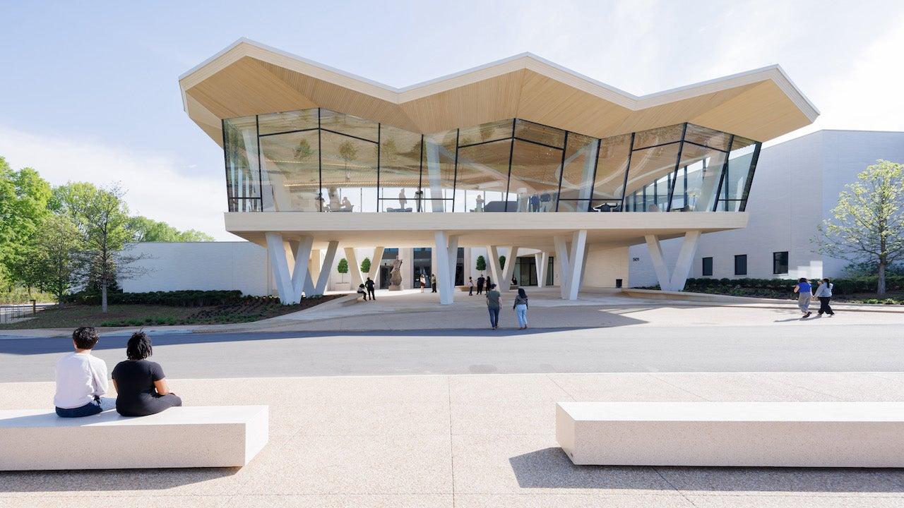 Futuristic building with angular roof and glass facade, people sitting and walking nearby.