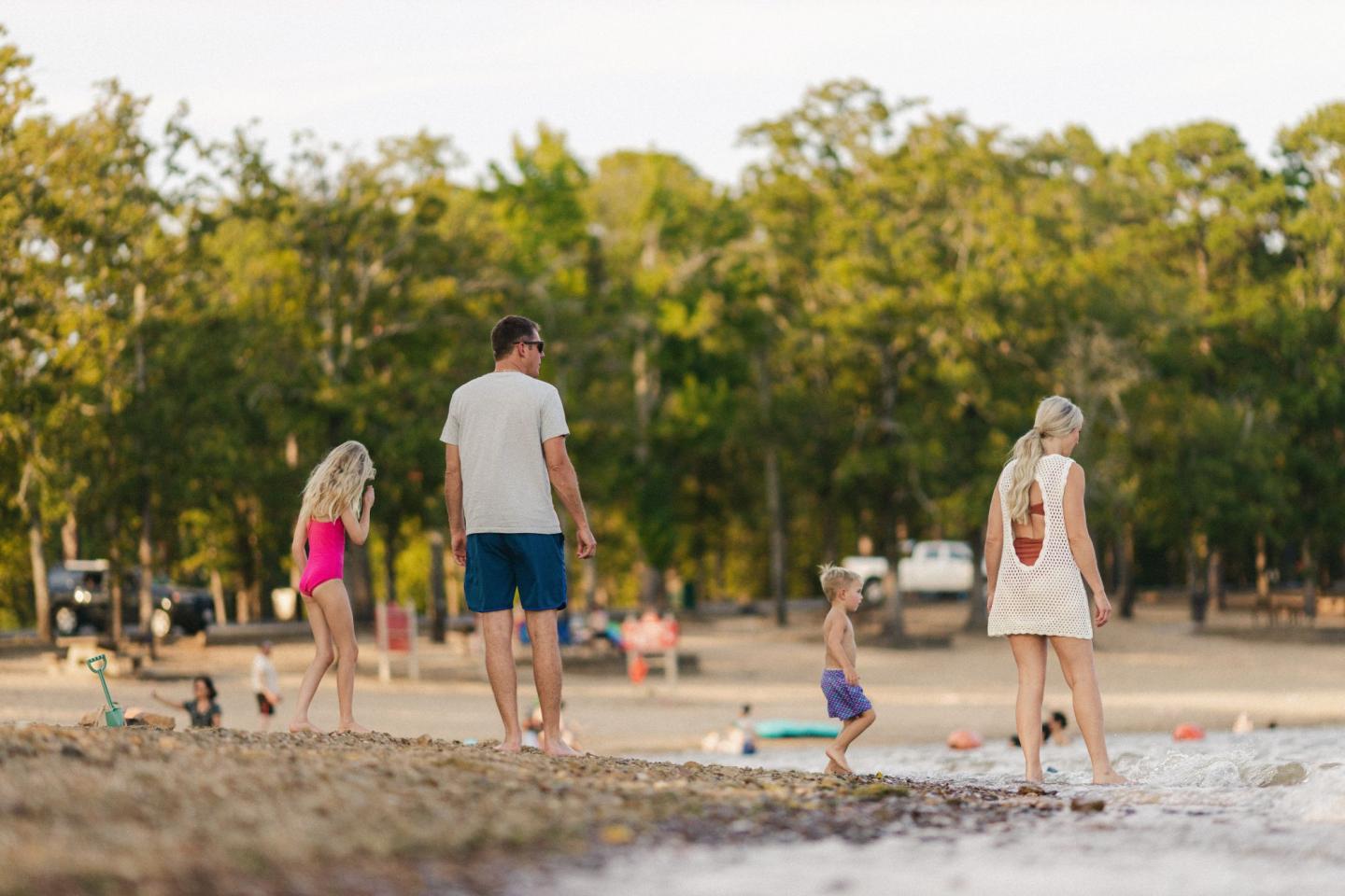 Family walking along a lakeside beach with trees in the background.