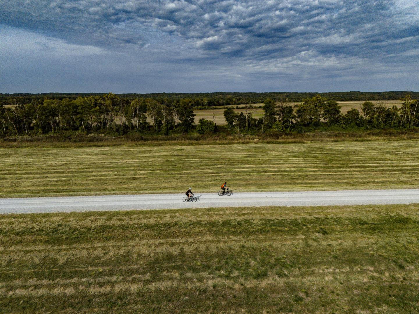 Two cyclists on a rural road, surrounded by fields and trees under a cloudy sky.