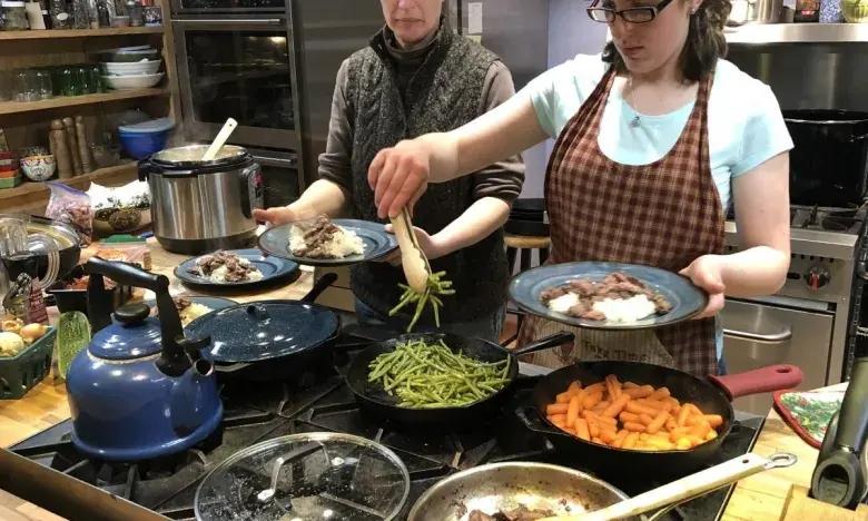 Two people in a kitchen plating meals with vegetables and meat.