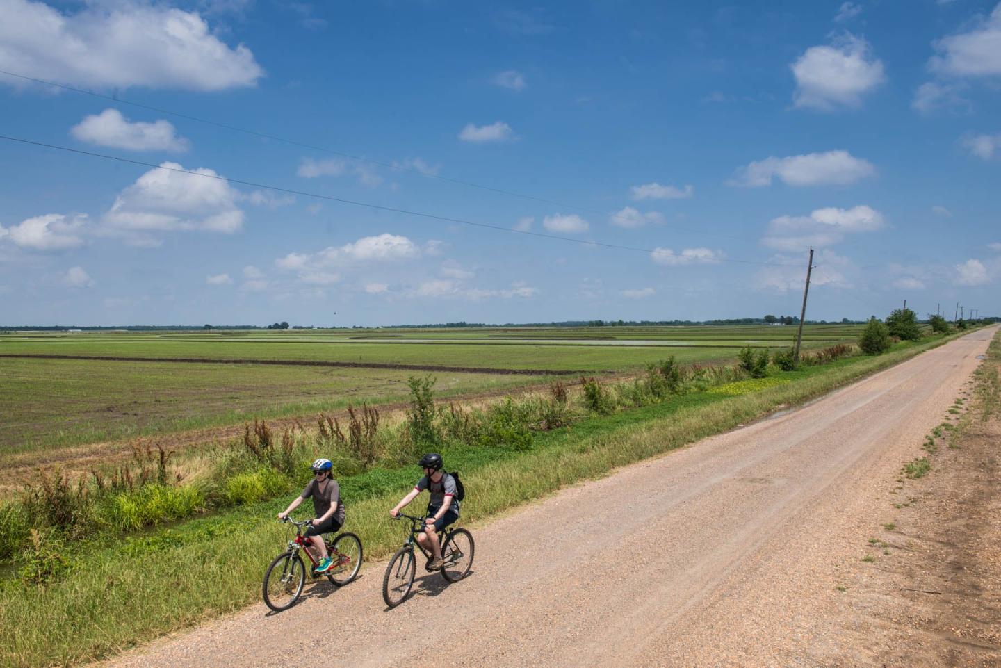 Cyclists ride on a rural dirt road under a clear blue sky.