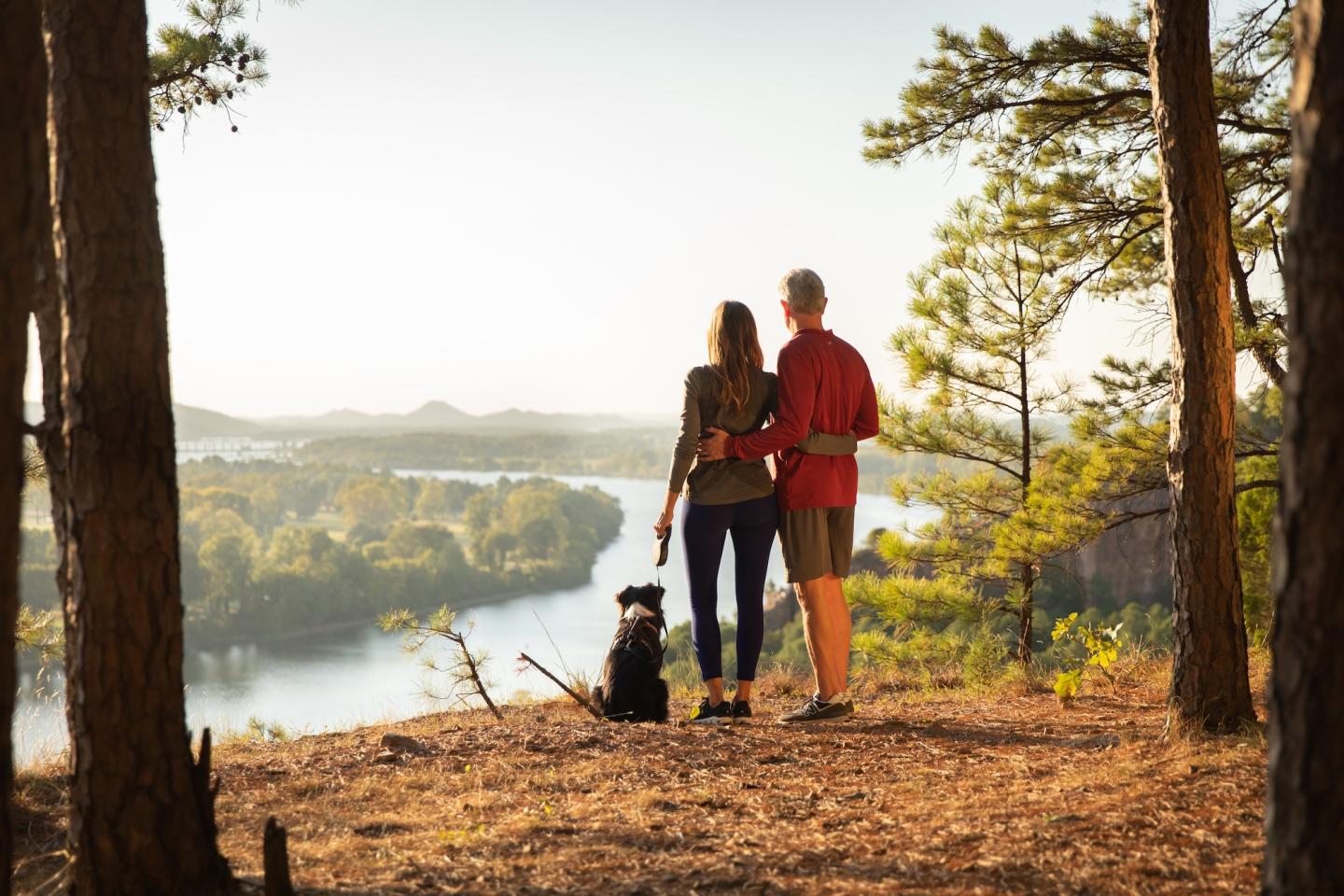 Couple and dog enjoy a scenic river view from a wooded hilltop.
