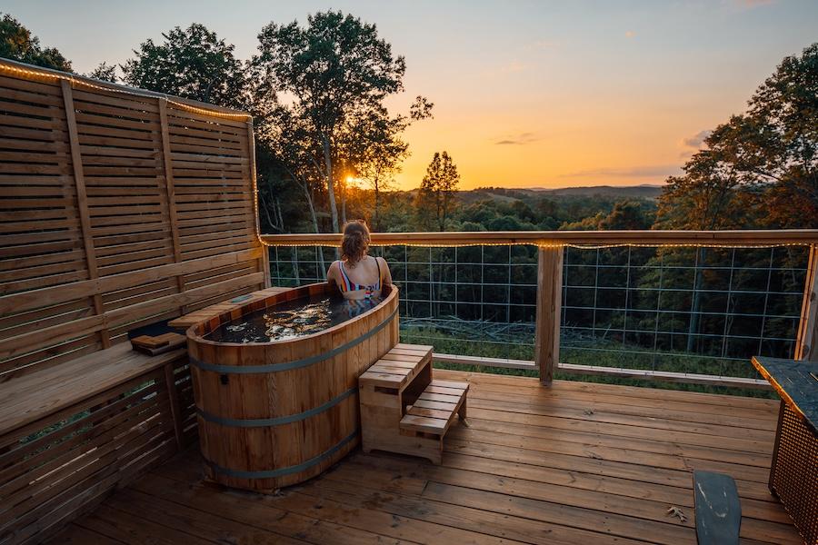 Woman relaxing in a wooden hot tub on a deck, overlooking a sunset view.